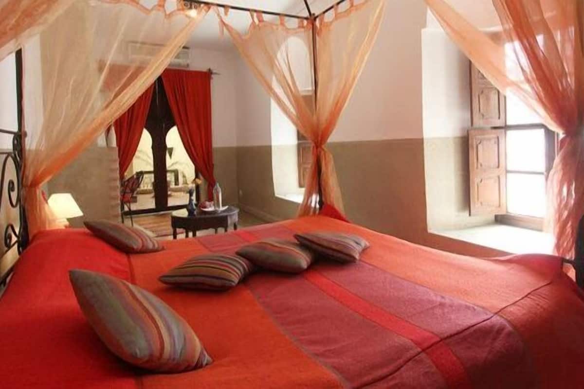 A bed with a canopy, a view of a room with a window, and a chair in the background.