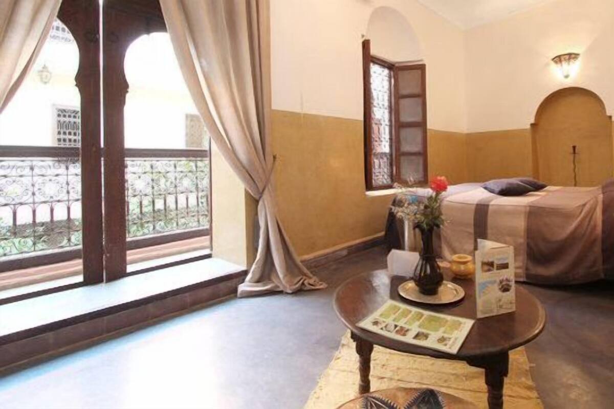 A room with a bed, a table with a magazine, a lamp, and a window with curtains.