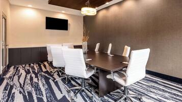 Un salón de conferencias con una mesa de madera oscura, sillas blancas, una alfombra estampada, un televisor montado y un jarrón decorativo.