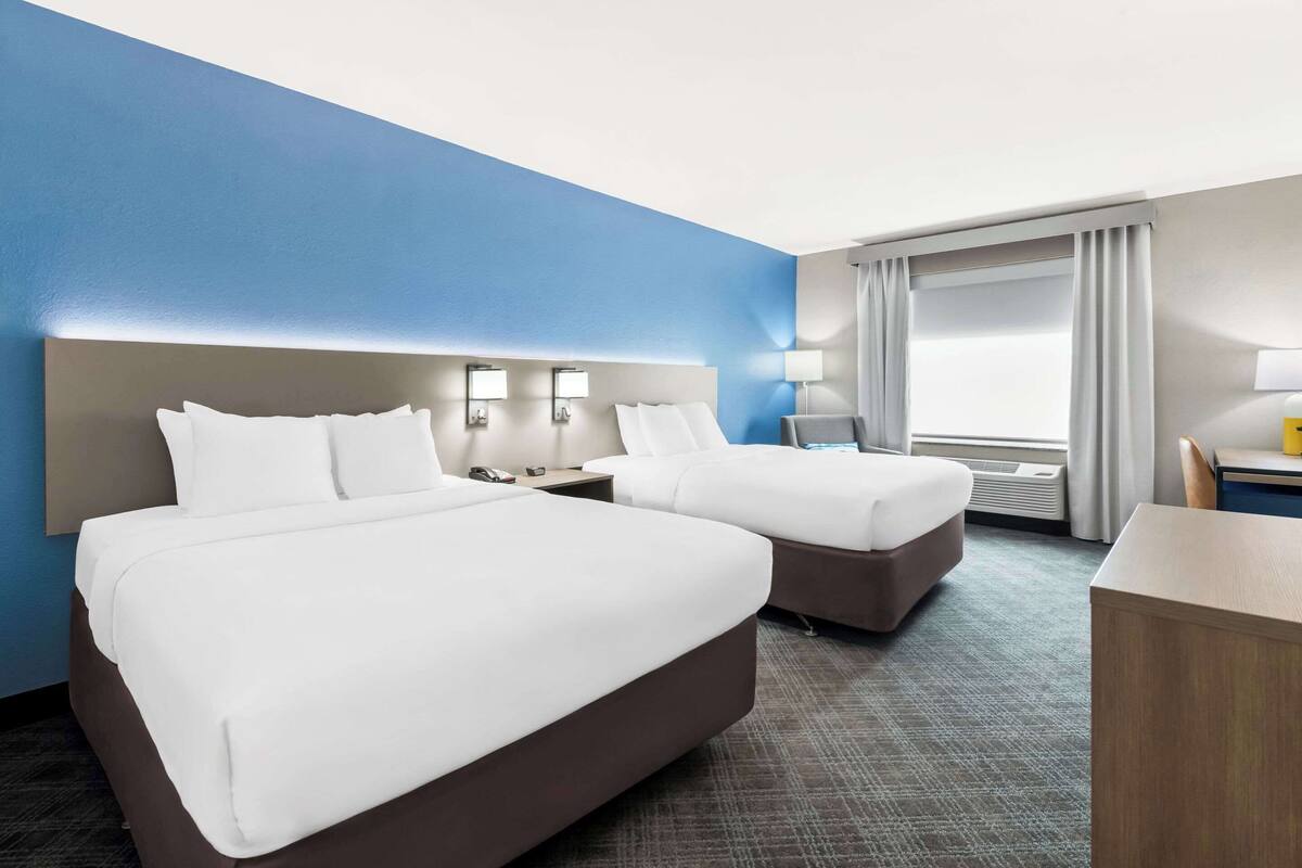 Une chambre d’hôtel avec deux lits, un bureau, une chaise, une lampe et une grande fenêtre avec des rideaux.