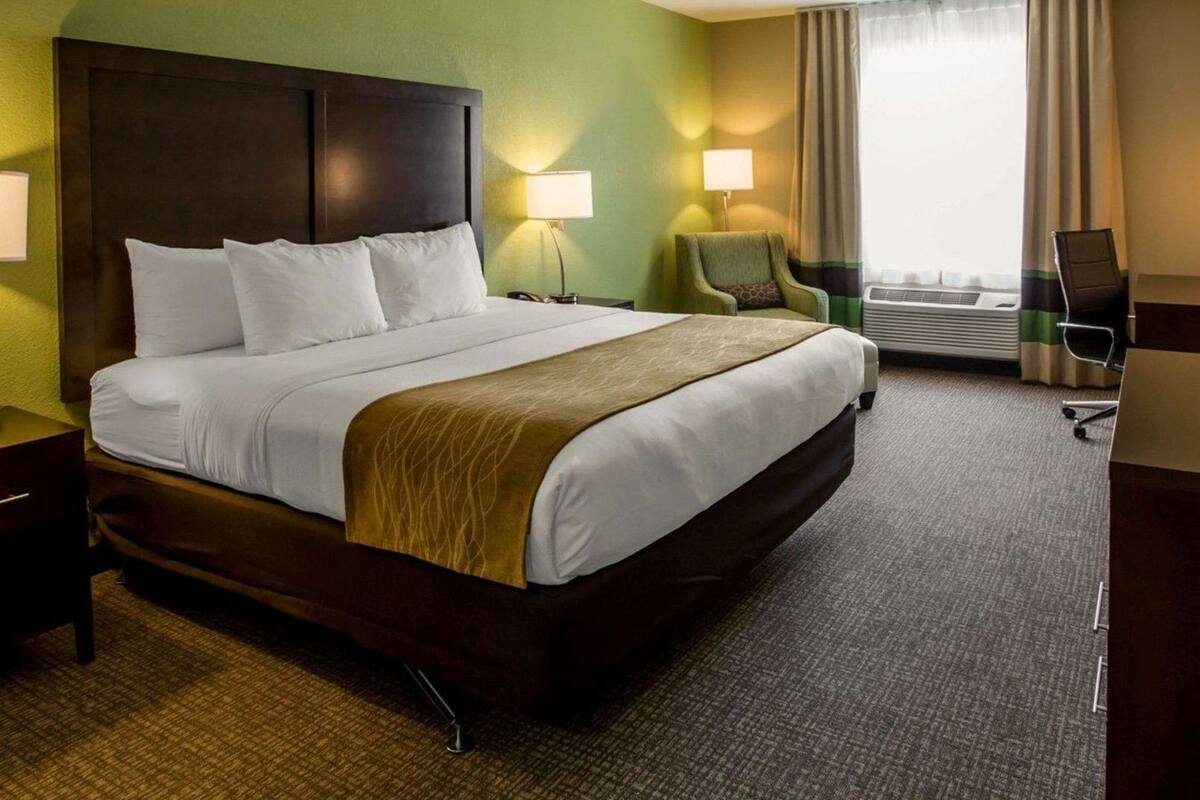 Une chambre d’hôtel avec un grand lit, un bureau, une chaise, une lampe et une fenêtre avec des rideaux.