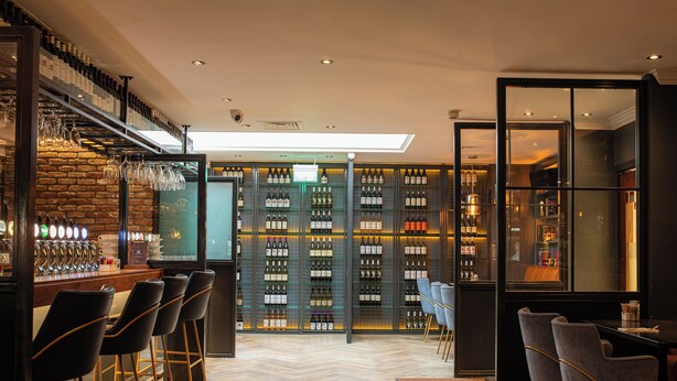Zona de bar con pared de ladrillo, varias botellas de vino y taburetes.