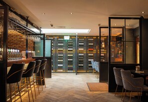 Zona de bar con pared de ladrillo, varias botellas de vino y taburetes.