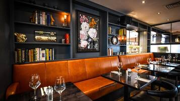 A restaurant with dark walls, a central seating area with a brown leather bench, and tables set with wine glasses and cutlery.