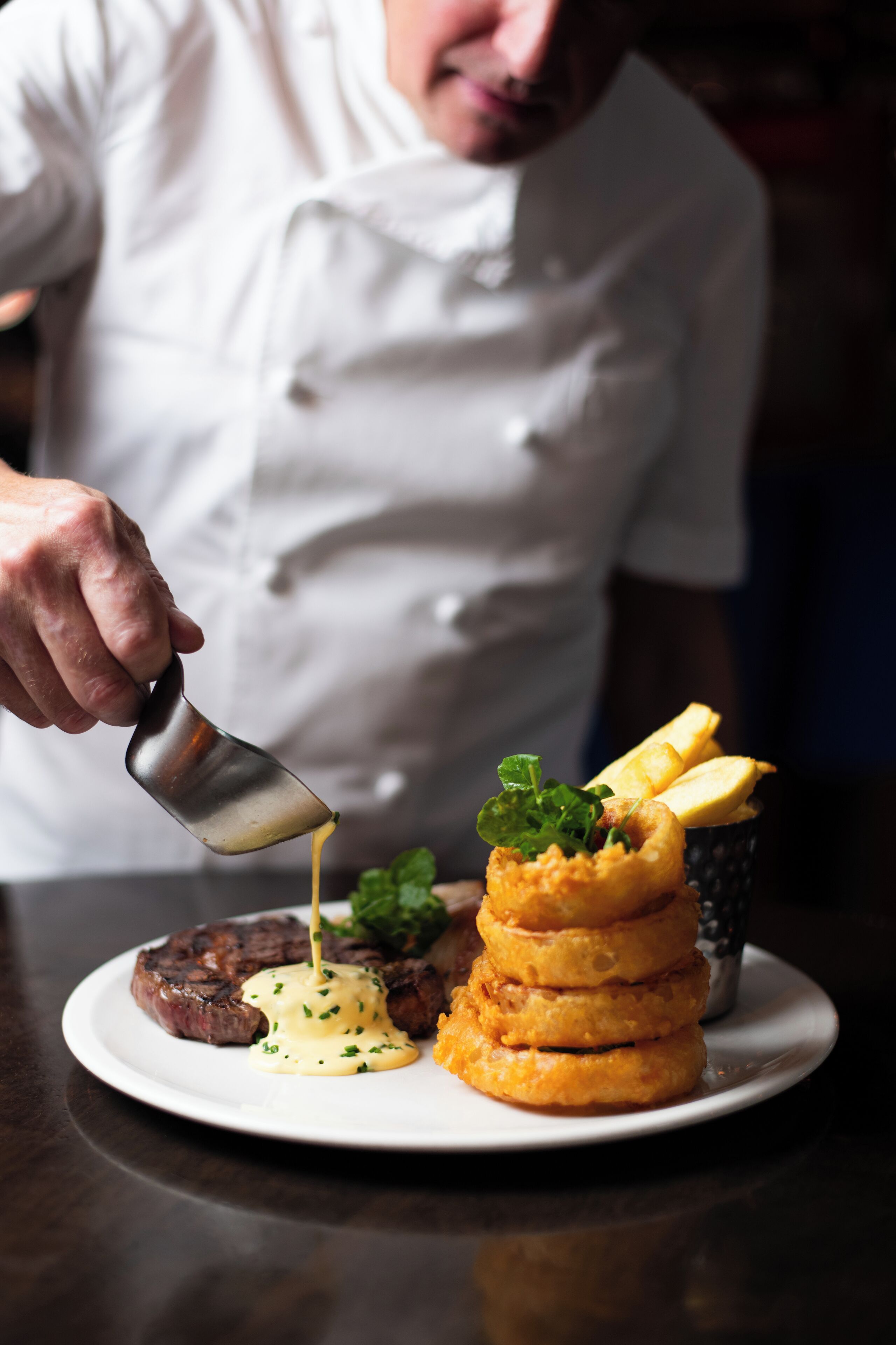 A chef pouring sauce onto a steak and onion rings on a plate.