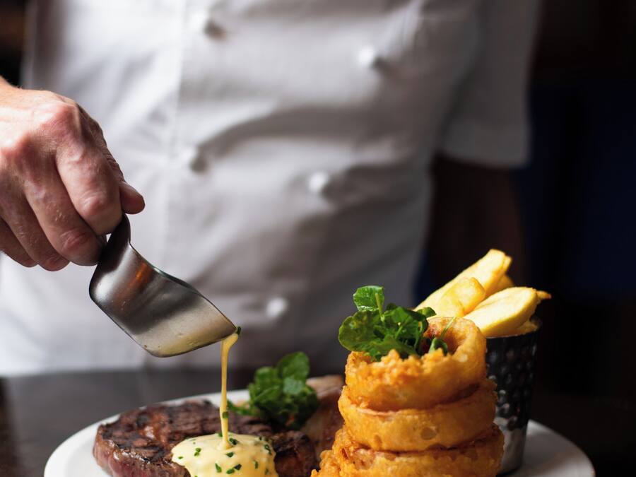 Un chef sirve salsa sobre un bistec y aros de cebolla en un plato.