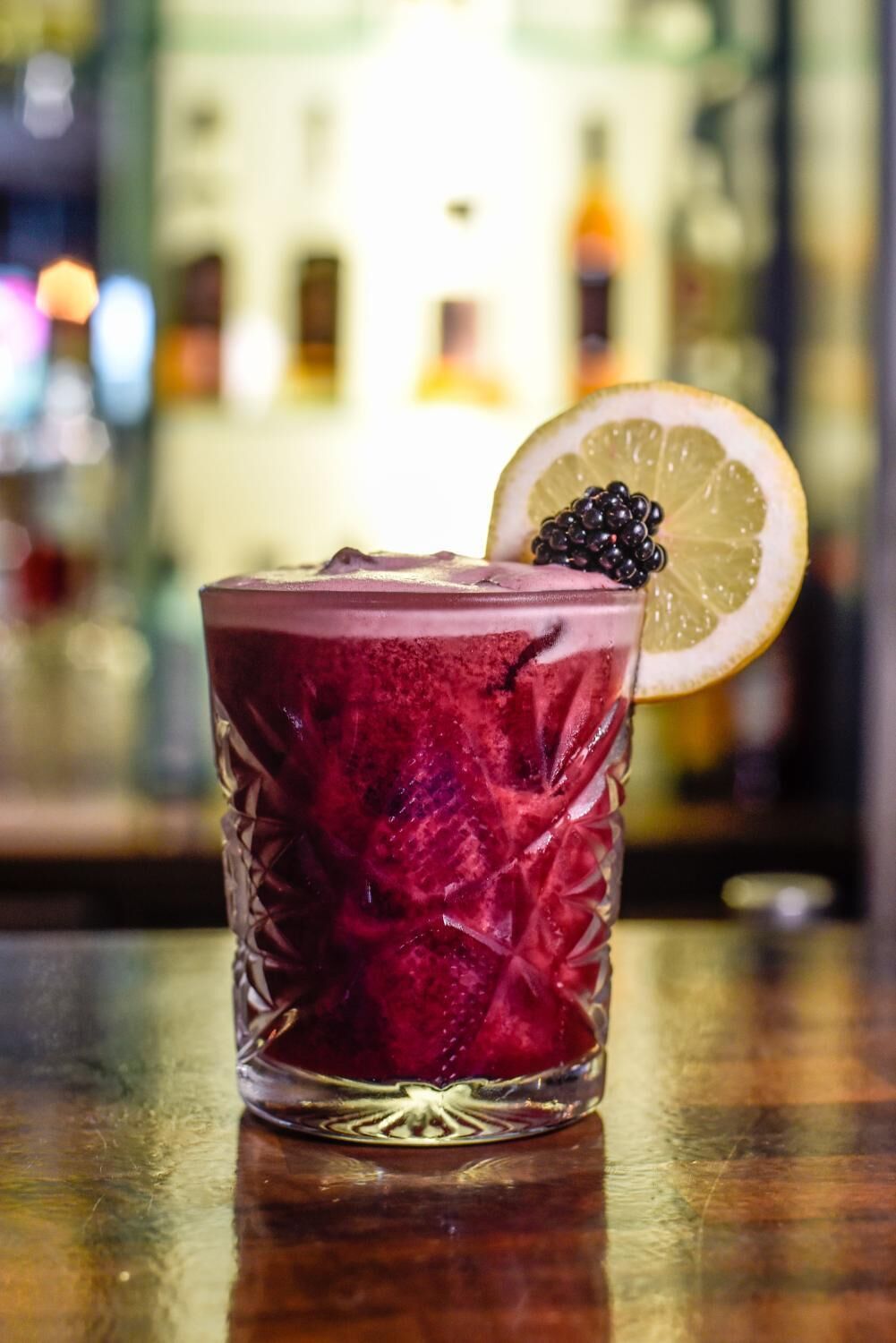 A glass of red beverage with a blackberry garnish on a bar counter.