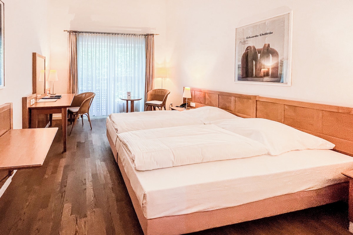 Ein Hotelzimmer mit einem Doppelbett, einem Schreibtisch mit Stuhl, einem kleinen Tisch ebenfalls mit Stuhl und einem großen Fenster mit Vorhängen.