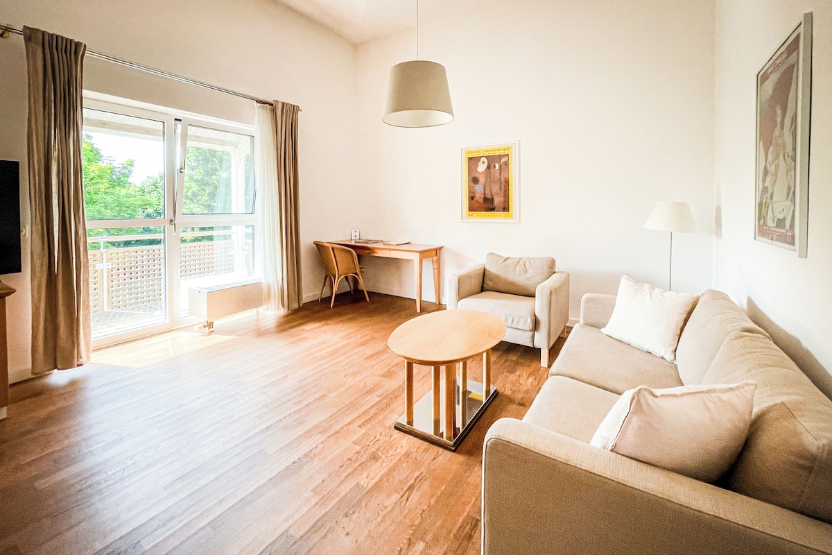 Ein geräumiges Wohnzimmer mit Holzboden, einem beigen Sofa, einem runden Couchtisch und einem Schreibtisch mit Stuhl. Ein großes Fenster mit Vorhängen, eine Deckenleuchte und ein gerahmtes Bild an der Wand sind ebenfalls vorhanden.