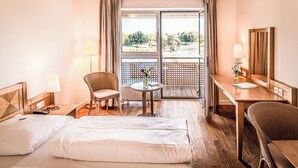 A hotel room with a bed, a desk, a chair, a small table with a vase, and a view of the outdoors through a sliding door. - Golfhotel & Restaurant Lindenhof (Bad Vilbel)