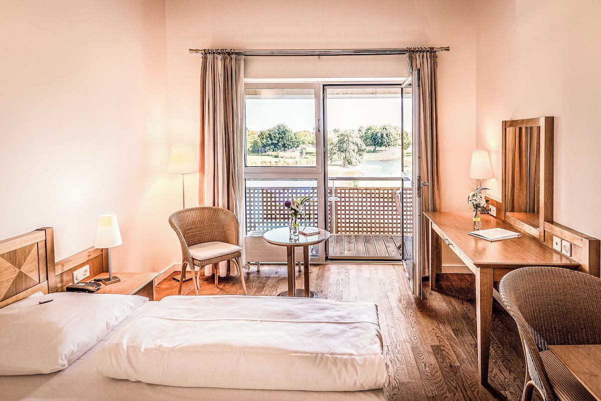 Ein Hotelzimmer mit Bett, Schreibtisch, Stuhl, einem kleinen Tisch mit einer Vase und Blick ins Freie durch eine Schiebetür.