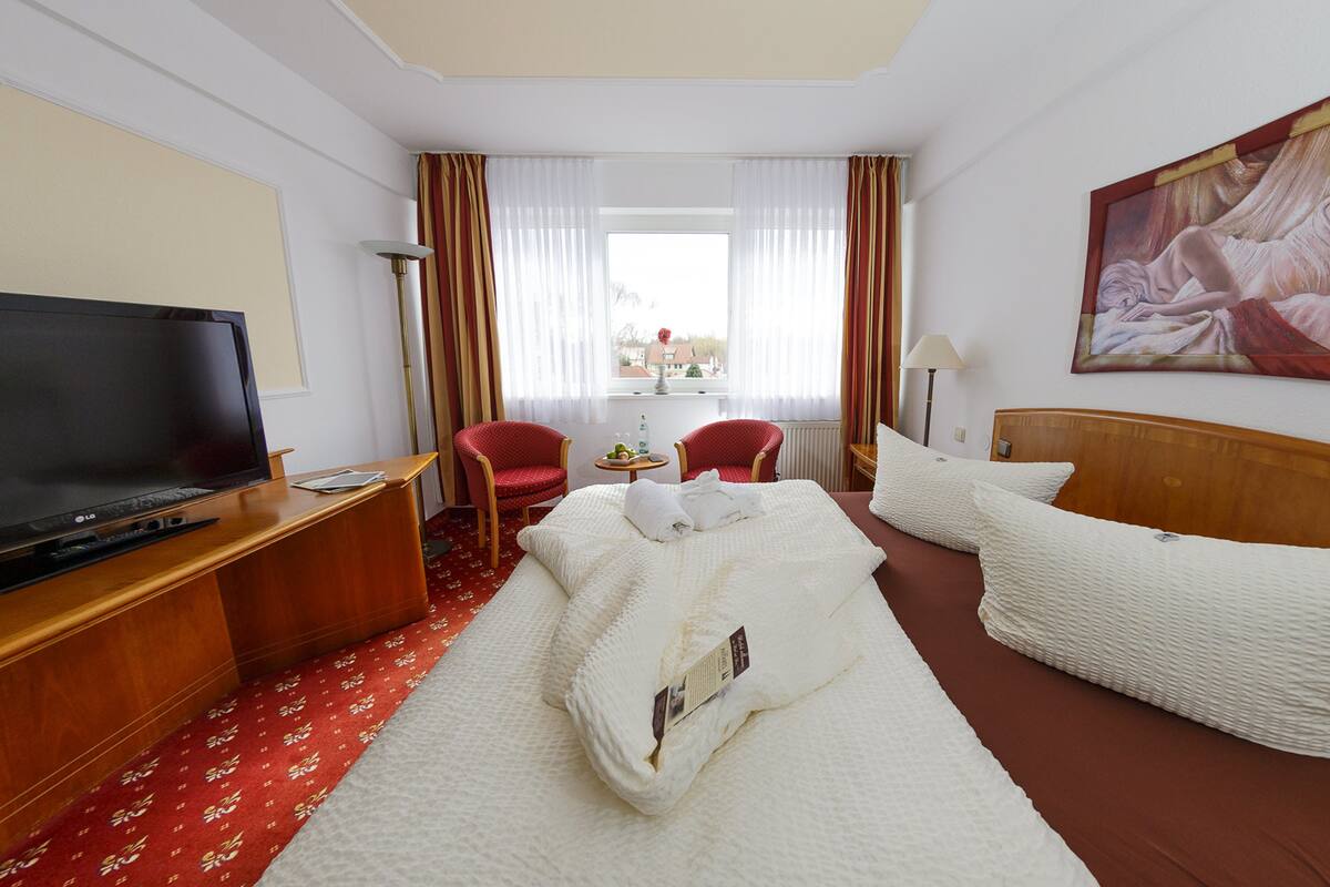 A hotel room with a bed, a desk with a TV, two chairs, a window with curtains, and a painting on the wall.