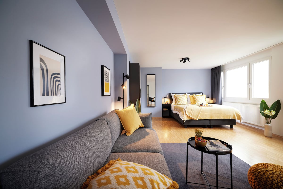 Een moderne slaapkamer met een grijze bank, een bed met geel beddengoed en een klein tafeltje met een plantje.