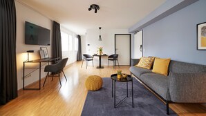 Een moderne woonkamer met een grijze bank, een rond geel poufje, een klein rond salontafeltje, een aan de muur bevestigde televisie, en een eethoek met een tafel en stoelen.