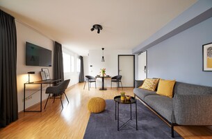 A modern living room with a grey sofa, a round yellow ottoman, a small round coffee table, a TV mounted on the wall, and a dining area with a table and chairs.