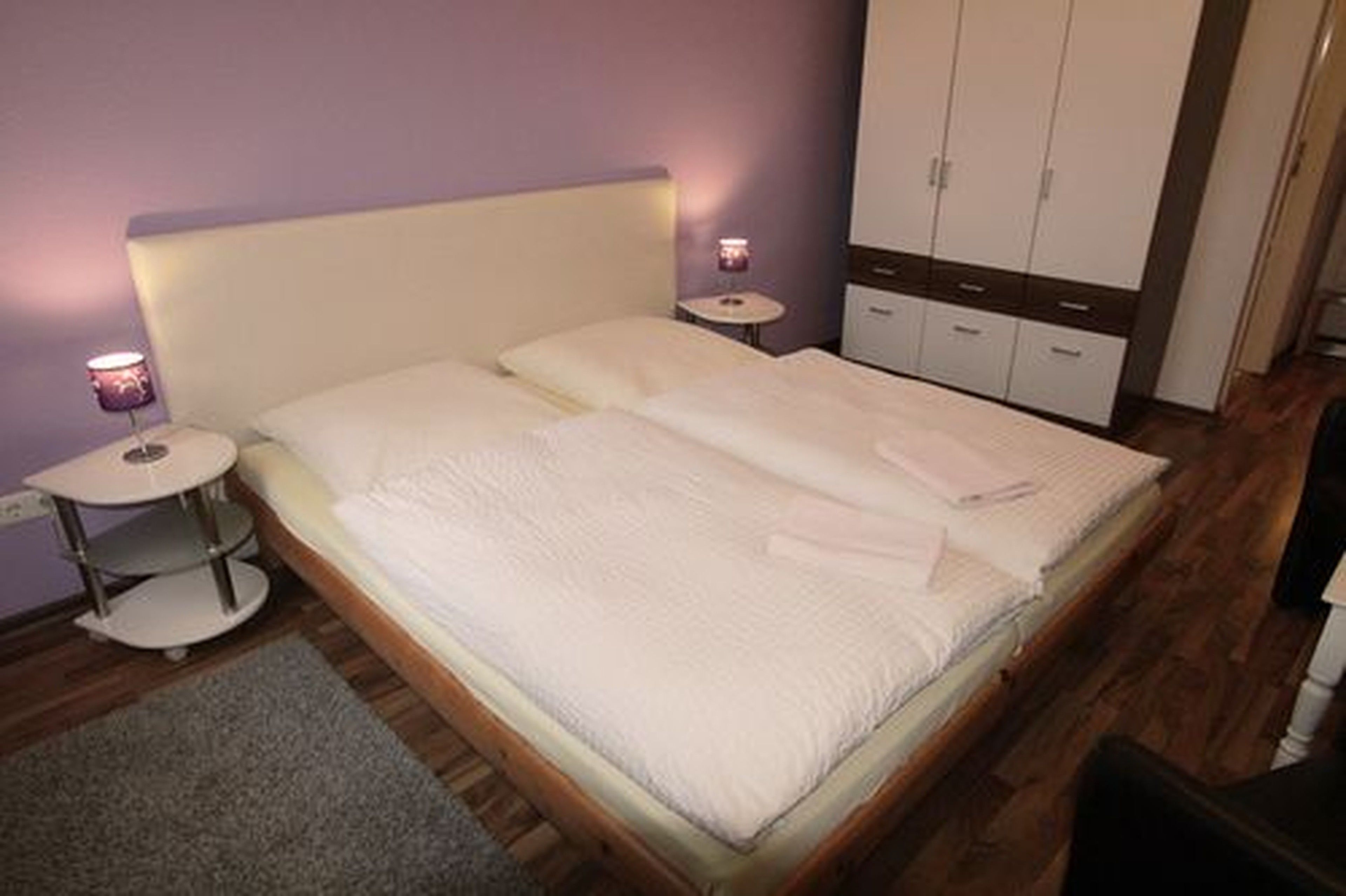 A neatly made bed with a headboard, a bedside table with a lamp, and a wardrobe in a room with wooden flooring.