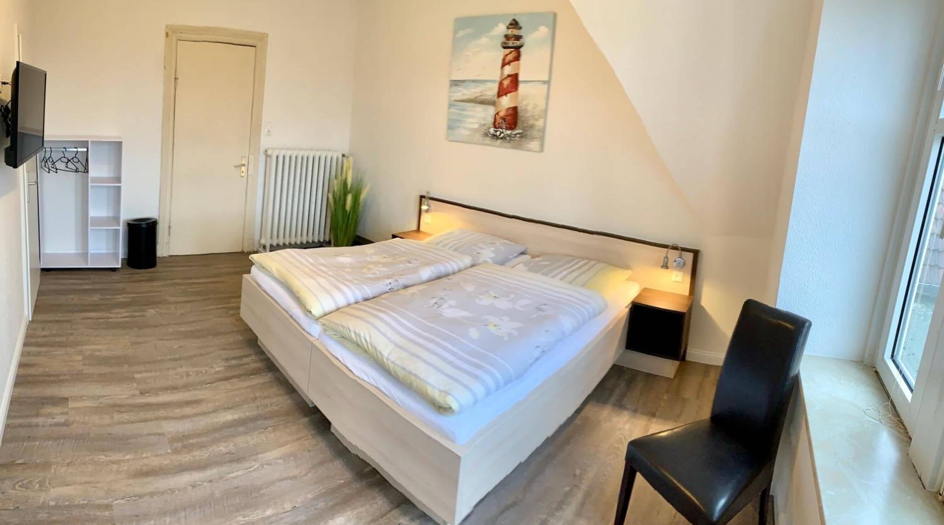 A bedroom with a bed, bedside tables, a chair, a radiator, a door, and a painting of a lighthouse.