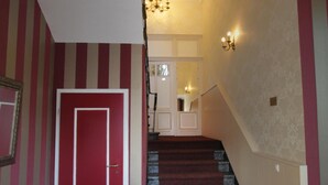 Una escalera que desemboca en un rellano con candelabro y apliques de pared, rematada con una puerta roja.