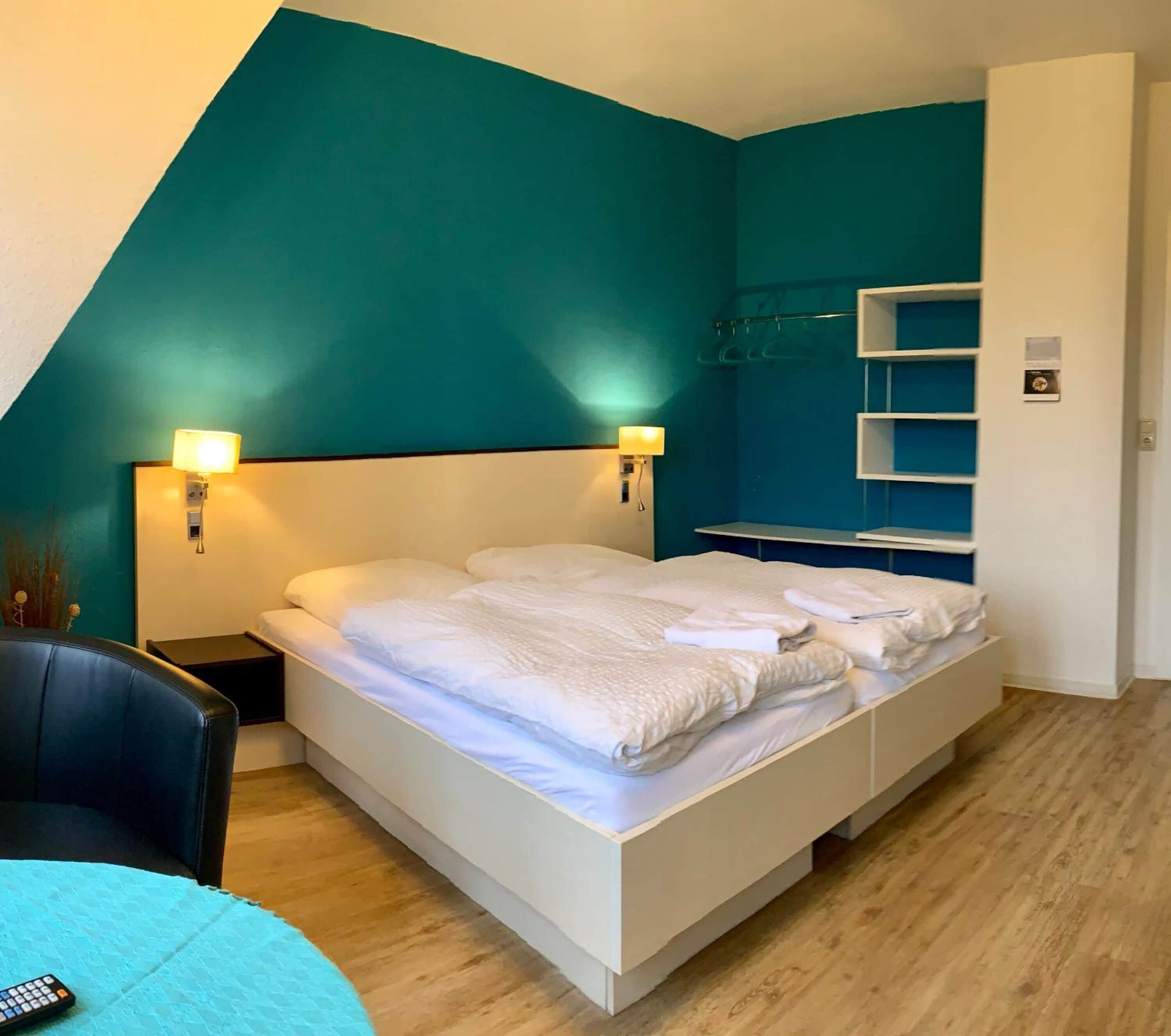 A modern bedroom with a white bed, teal walls, and a wooden floor. There is a nightstand with a lamp, a chair, and a small table with a remote control.
