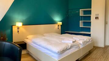 Quarto moderno com cama branca, paredes verde-água e piso de madeira. Há uma mesinha de cabeceira com abajur, uma cadeira e uma pequena mesa com um controle remoto.