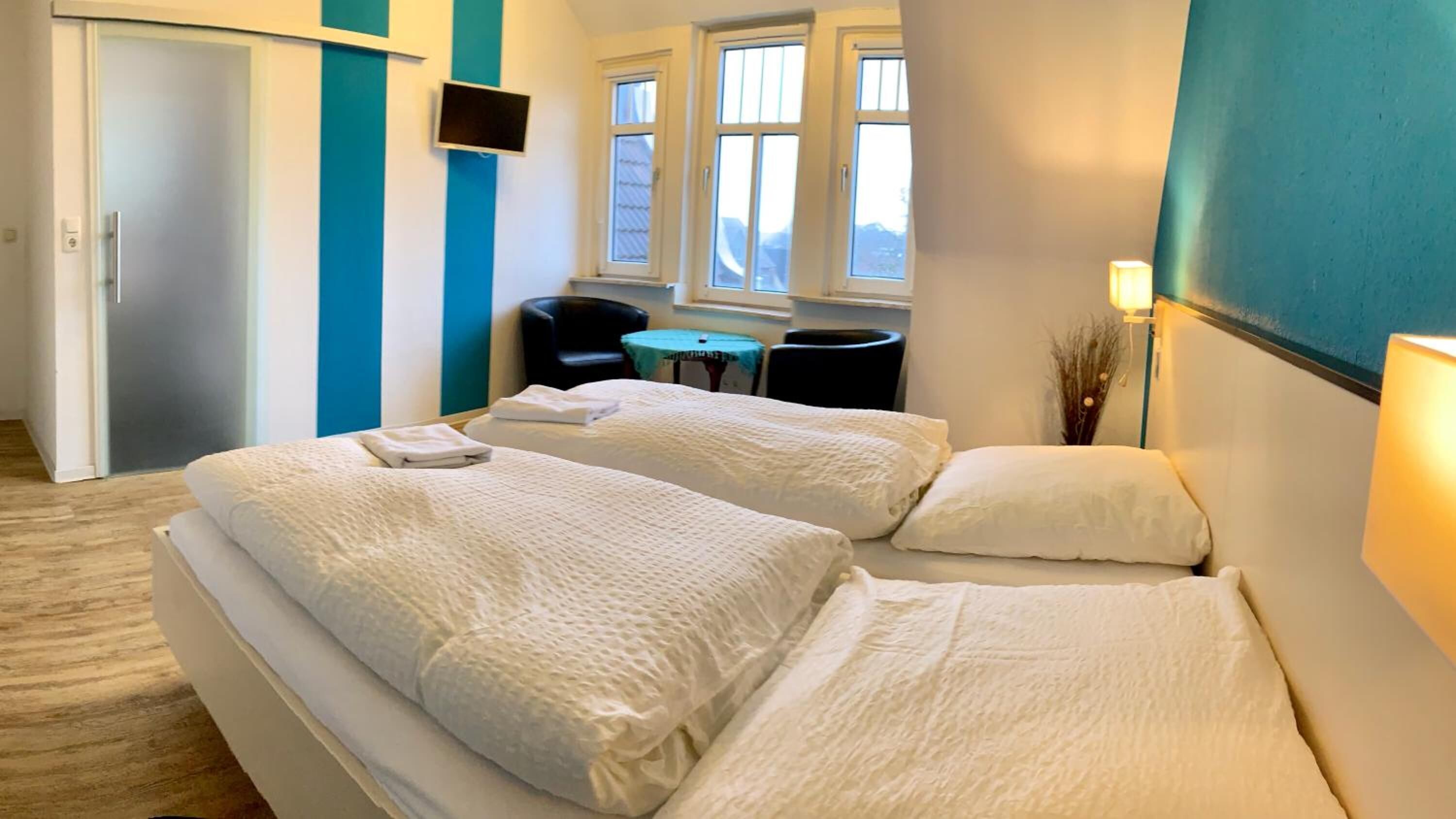 A hotel room with two beds, a TV, a chair, a small table, and windows with a view.