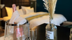 Een hotelkamer met een bed, een nachtkastje met een fles The Duke Gin, een glas en een vaas met gedroogde bloemen.