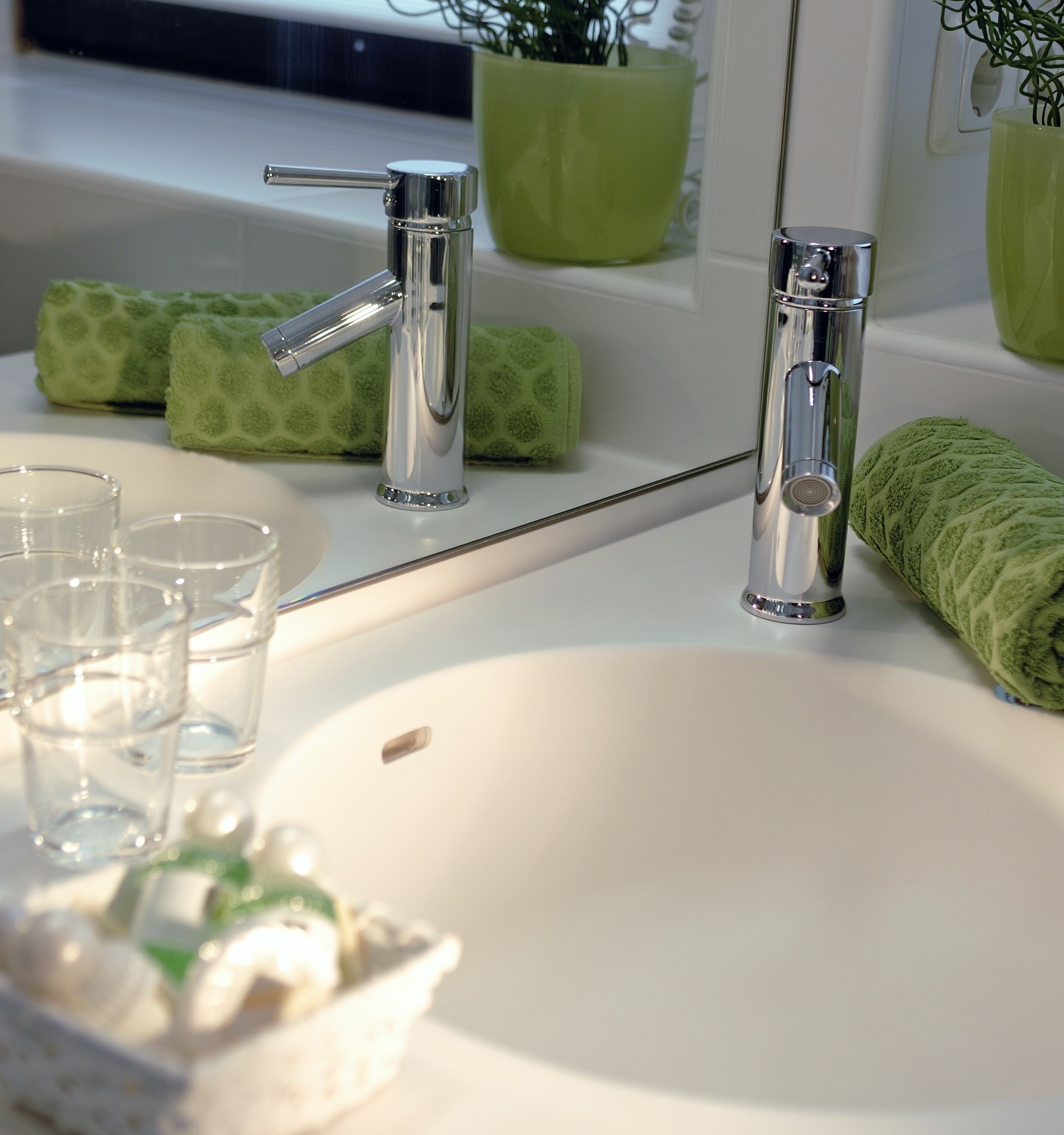 A bathroom with a white sink, two chrome faucets, and a mirror. There are green towels and a green plant in a vase on the counter.