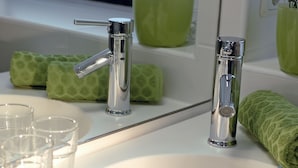 Ein Badezimmer mit einem weißen Waschbecken, zwei Chromarmaturen und einem Spiegel. Auf dem Waschtisch liegen grüne Handtücher und eine grüne Pflanze im Blumenvasen.