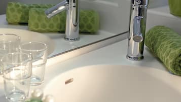 A bathroom with a white sink, two chrome faucets, and a mirror. There are green towels and a green plant in a vase on the counter.