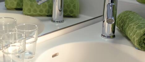 A bathroom with a white sink, two chrome faucets, and a mirror. There are green towels and a green plant in a vase on the counter.
