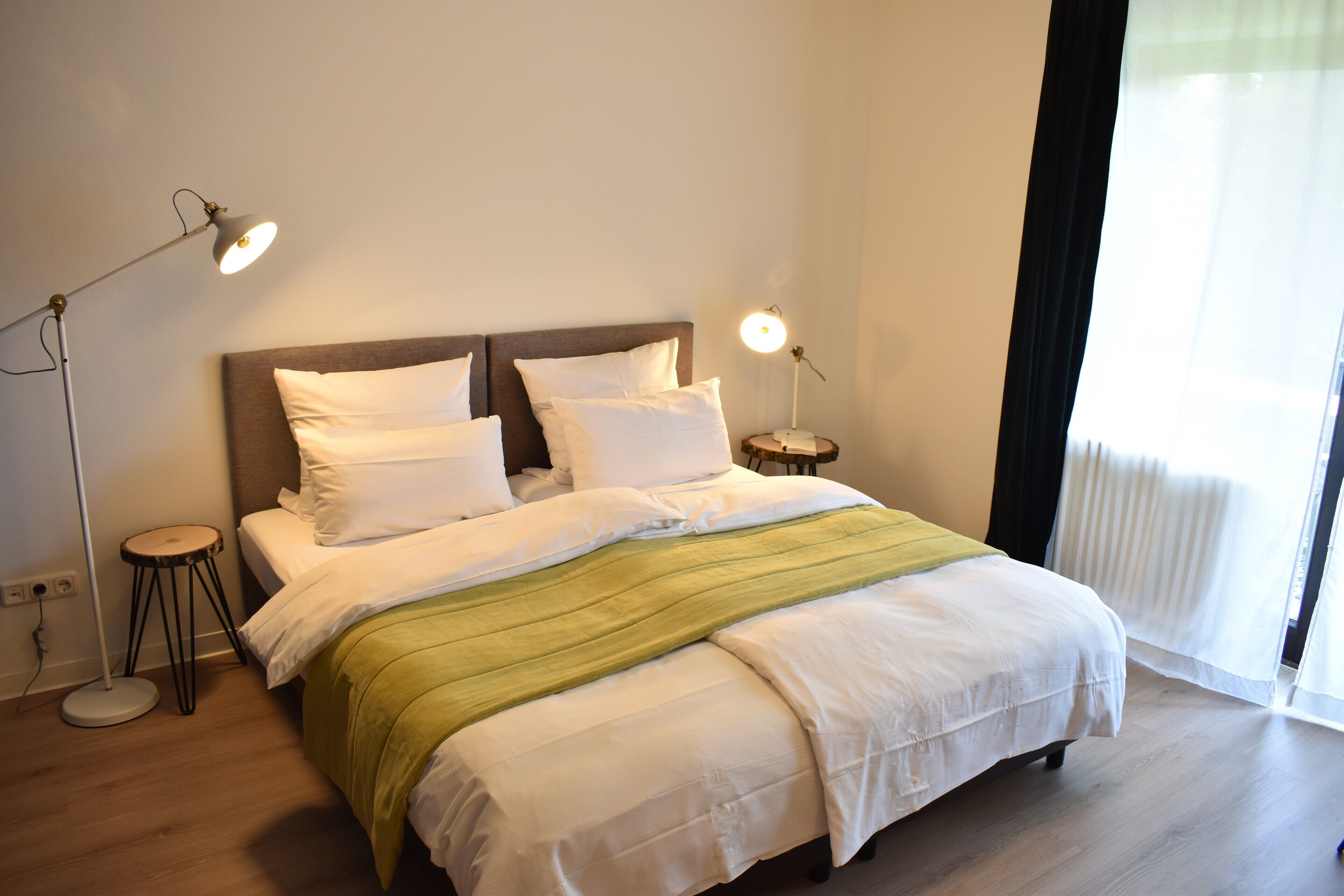 Een netjes opgemaakt bed met witte lakens en een groene plaid, twee bedlampjes en een raam met dunne gordijnen.