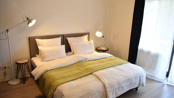 Een netjes opgemaakt bed met witte lakens en een groene plaid, twee bedlampjes en een raam met dunne gordijnen.
