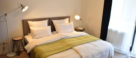 A neatly made bed with white linens and a green throw blanket, two bedside lamps, and a window with sheer curtains.