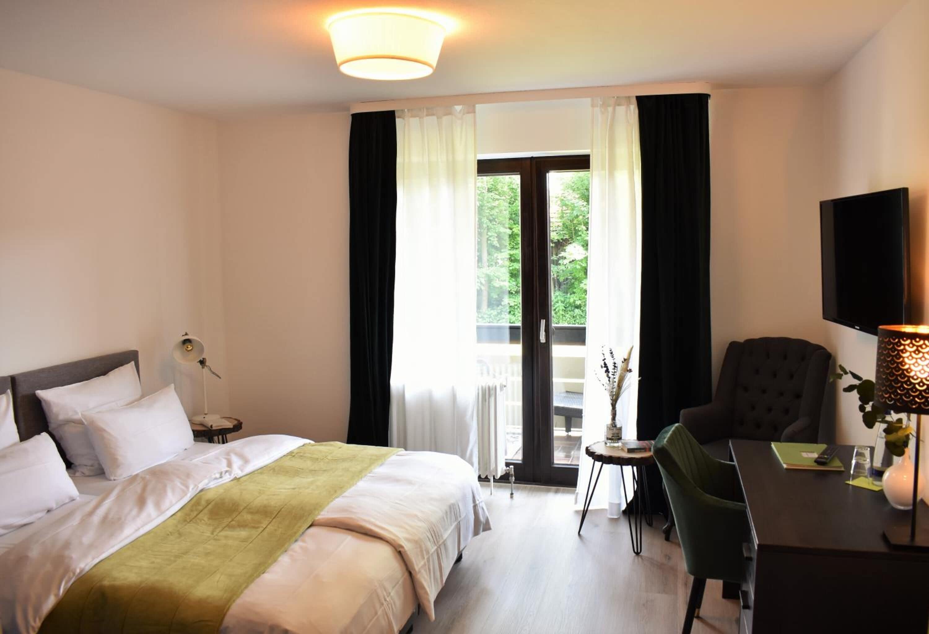 Hotellihuone, jossa on sänky, työpöytä, tuoli, lamppu ja ikkunasta avautuva näkymä vehreään maisemaan.