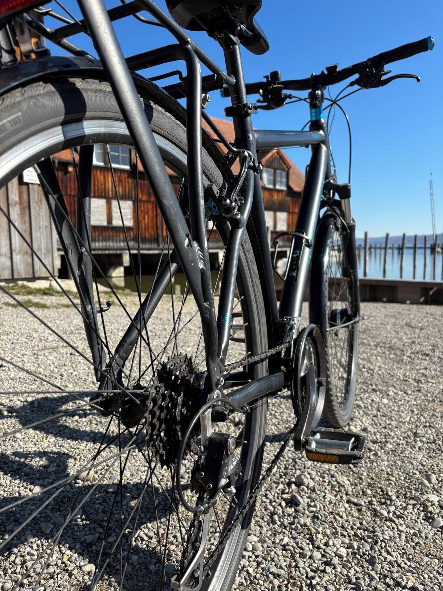Una bicicleta estacionada sobre grava, con un edificio de madera y una masa de agua al fondo.