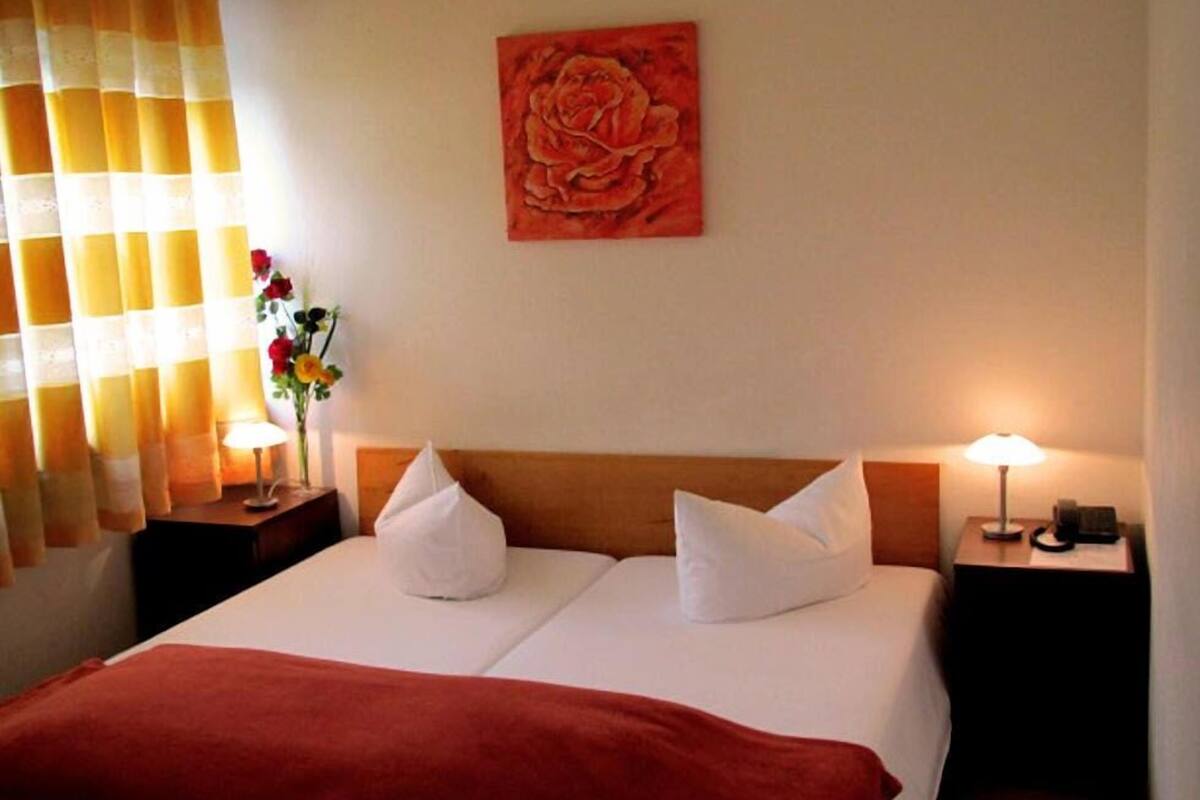 Ett hotellrum med en säng, två sängbord, en lampa, en vas med blommor och en tavla på väggen.