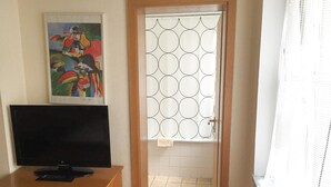 A room with a TV on a wooden stand, a colorful painting on the wall, and a window with a geometric pattern. - Pension Mona Lisa (Munich)