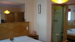 A hotel room with a bed, a bedside table with a lamp, a mirror, and a shower area. - Pension Mona Lisa (Munich)