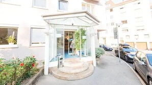 A hotel entrance with a glass canopy, a small garden with red flowers, and parked cars along the street. - Hotel Am Friedensplatz (Stuttgart)