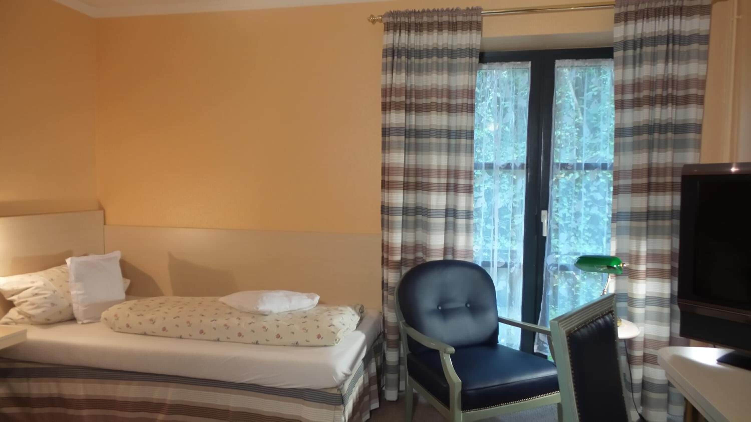 A hotel room with a bed, a chair, a TV, and a window with curtains.