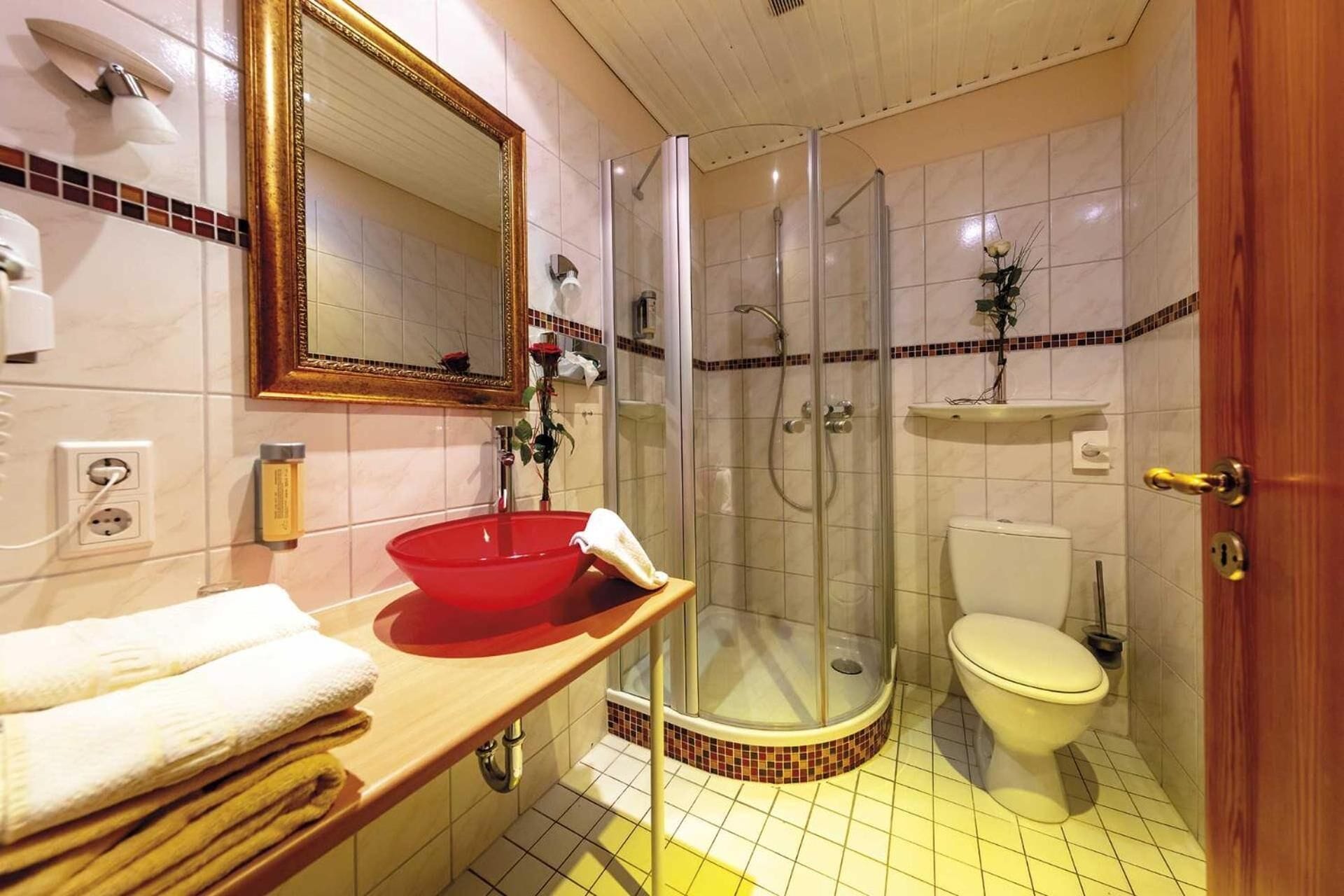 Un baño con un lavabo rojo, una ducha con mampara de vidrio, un inodoro y un tocador de madera con toallas.