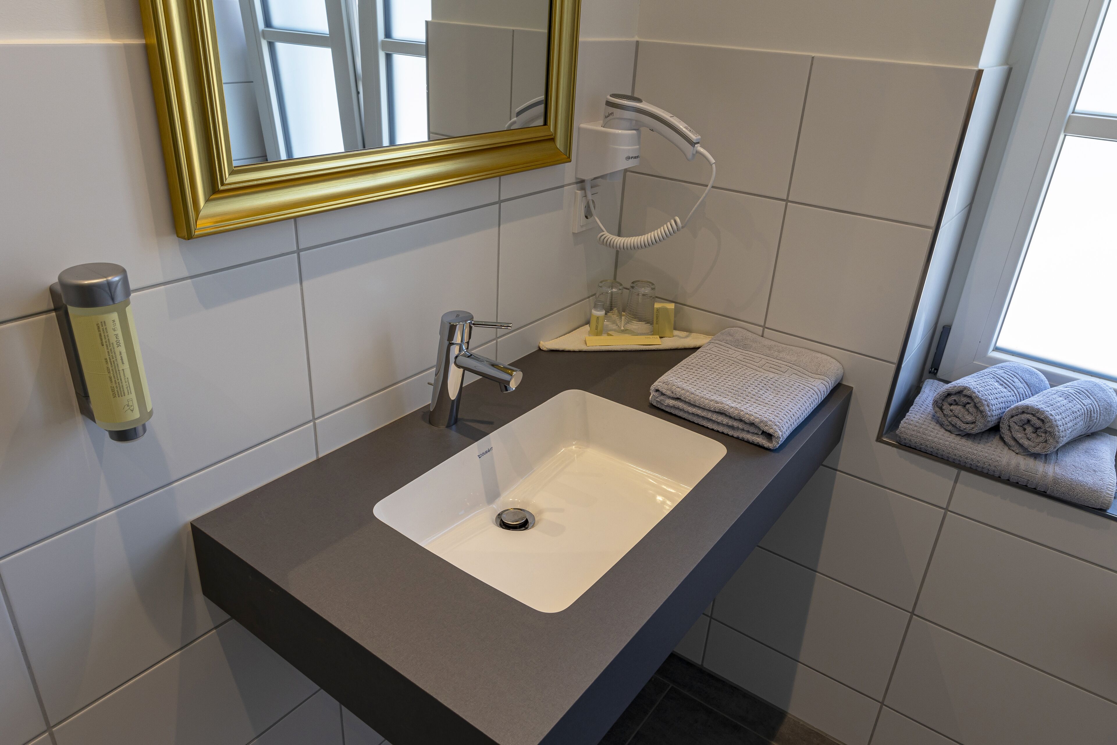 A bathroom with a white sink, a mirror with a gold frame, a towel rack, and a window with towels.