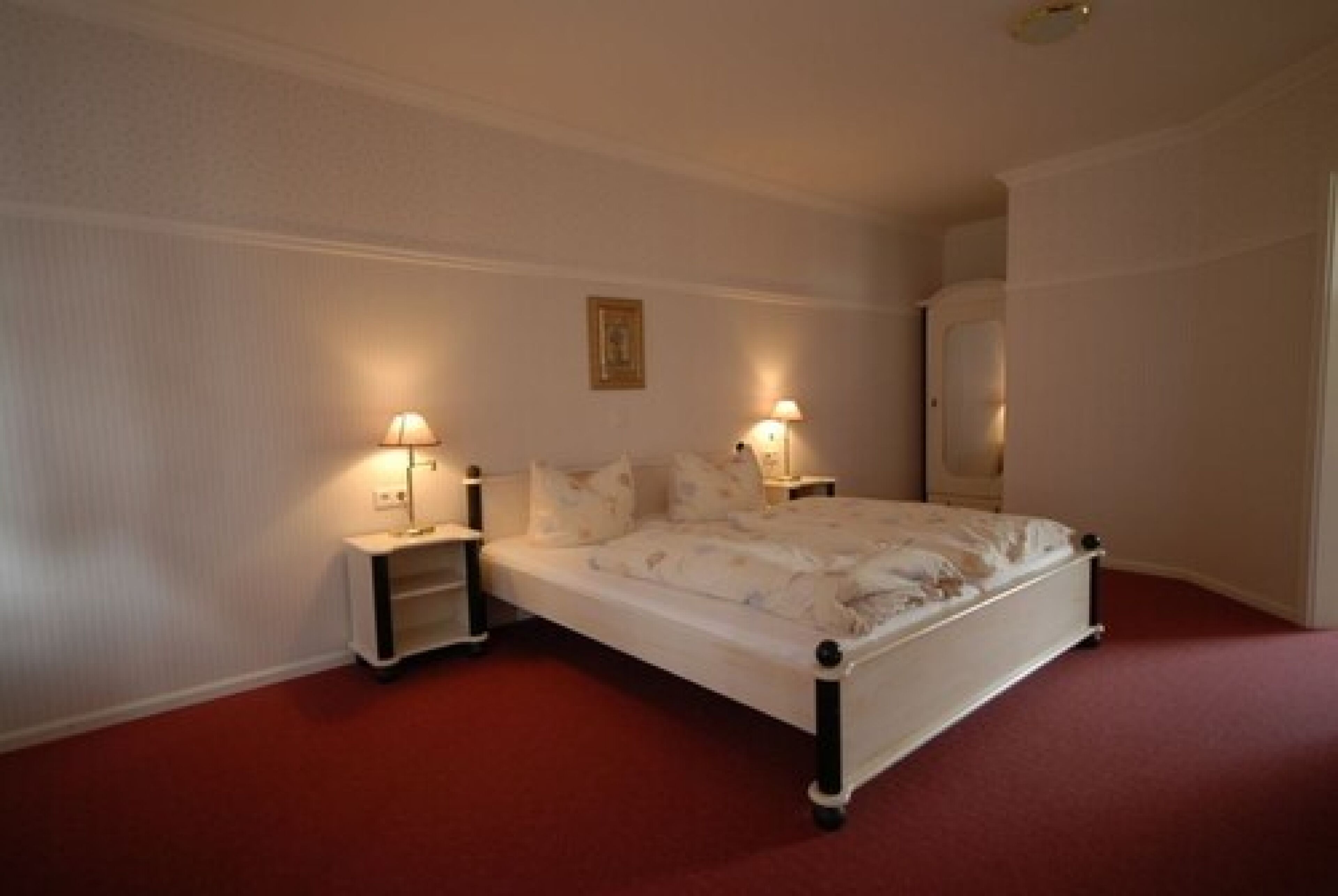 Un dormitorio con cama blanca, dos mesitas de noche con lámparas, alfombra roja y pared blanca con un cuadro enmarcado.