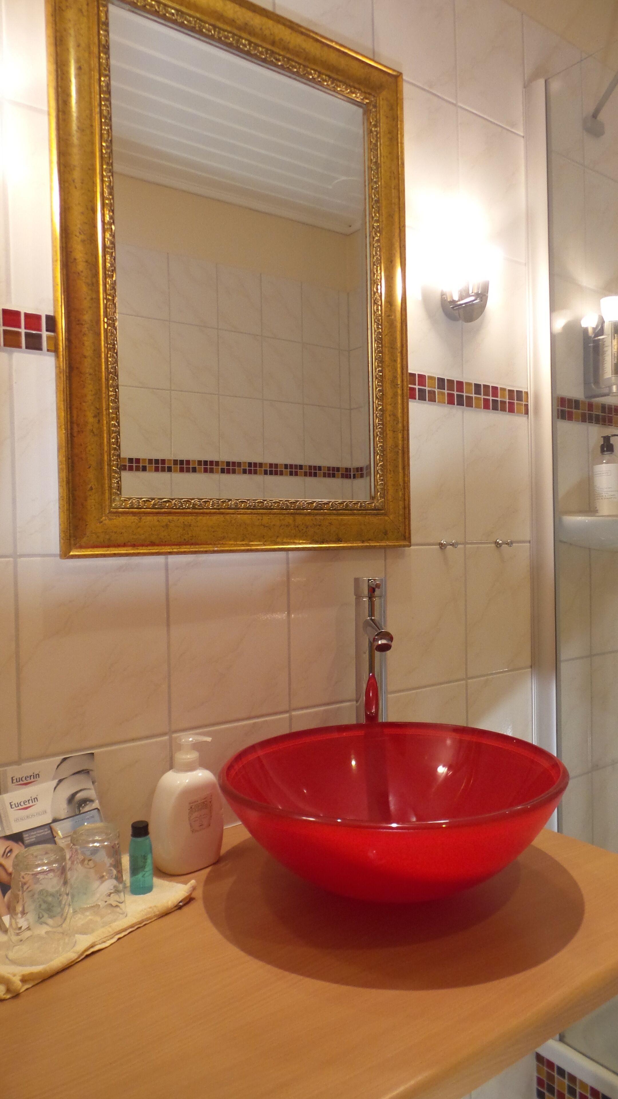 A bathroom with a red glass bowl sink, a gold-framed mirror, and toiletries on the counter.