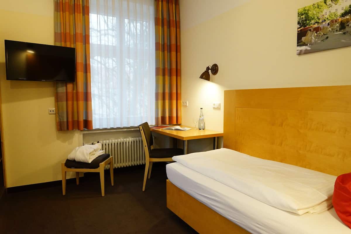 Una habitación de hotel con cama, escritorio, silla, radiador, televisor y ventana con cortinas.