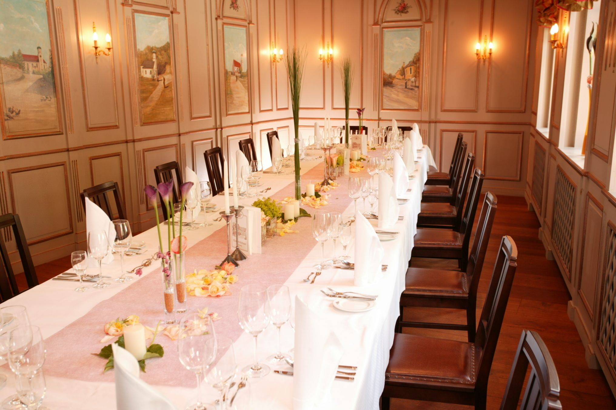 Una larga mesa de comedor dispuesta con manteles blancos, velas y centros de mesa florales en una habitación con paneles de madera y luces empotradas.