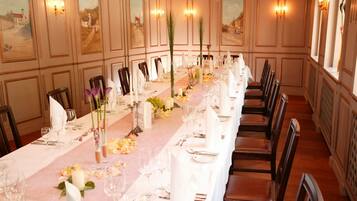 Una larga mesa de comedor dispuesta con manteles blancos, velas y centros de mesa florales en una habitación con paneles de madera y luces empotradas.