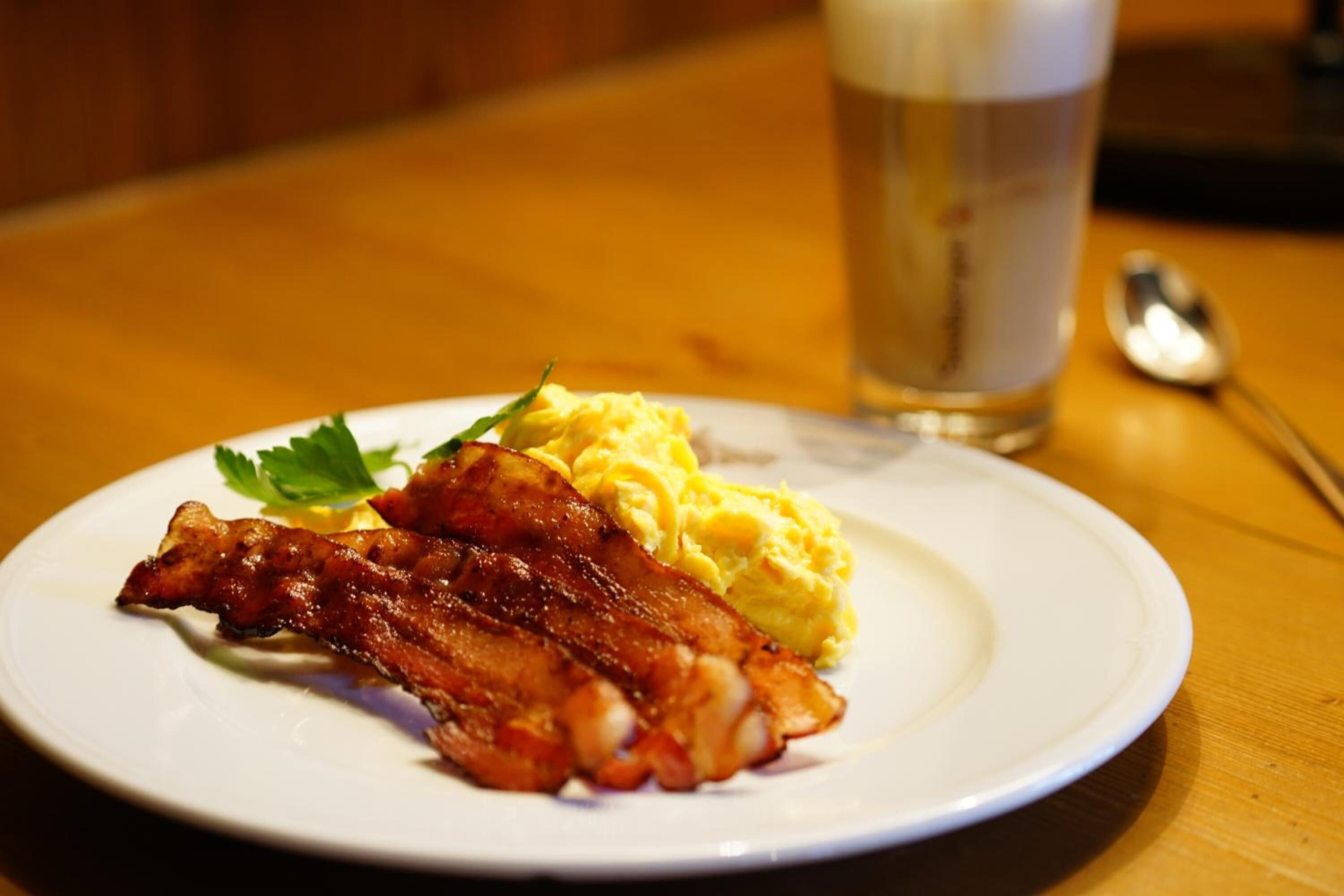 Ein Teller mit Speck und Rührei, ein Bierglas und ein Löffel auf einem Holztisch.