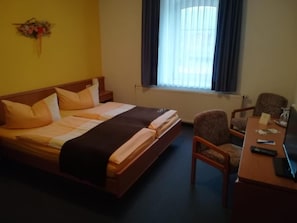 Une chambre d’hôtel comprenant un lit, un bureau avec une chaise, une fenêtre avec des rideaux et une petite table avec une télécommande.