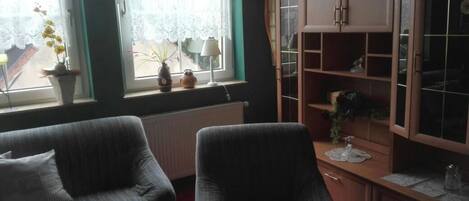 A living room with two armchairs, a wooden cabinet with glass doors, and a window with lace curtains.
